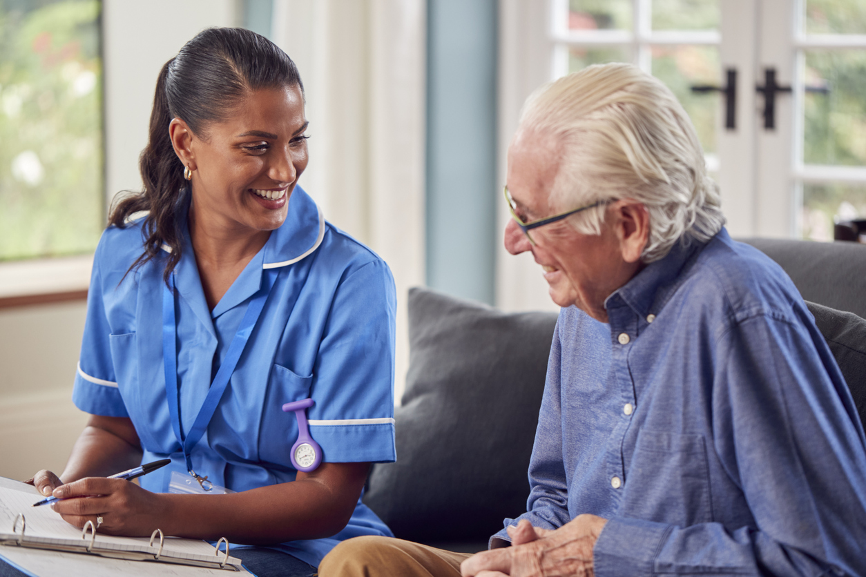 Senior Man At Home Talking To Female Nurse Or Care Worker